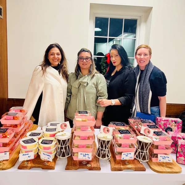 Four women stand behind a table laden with Monica's Mithai Cafe & Bar's Indian sweets and puddings.