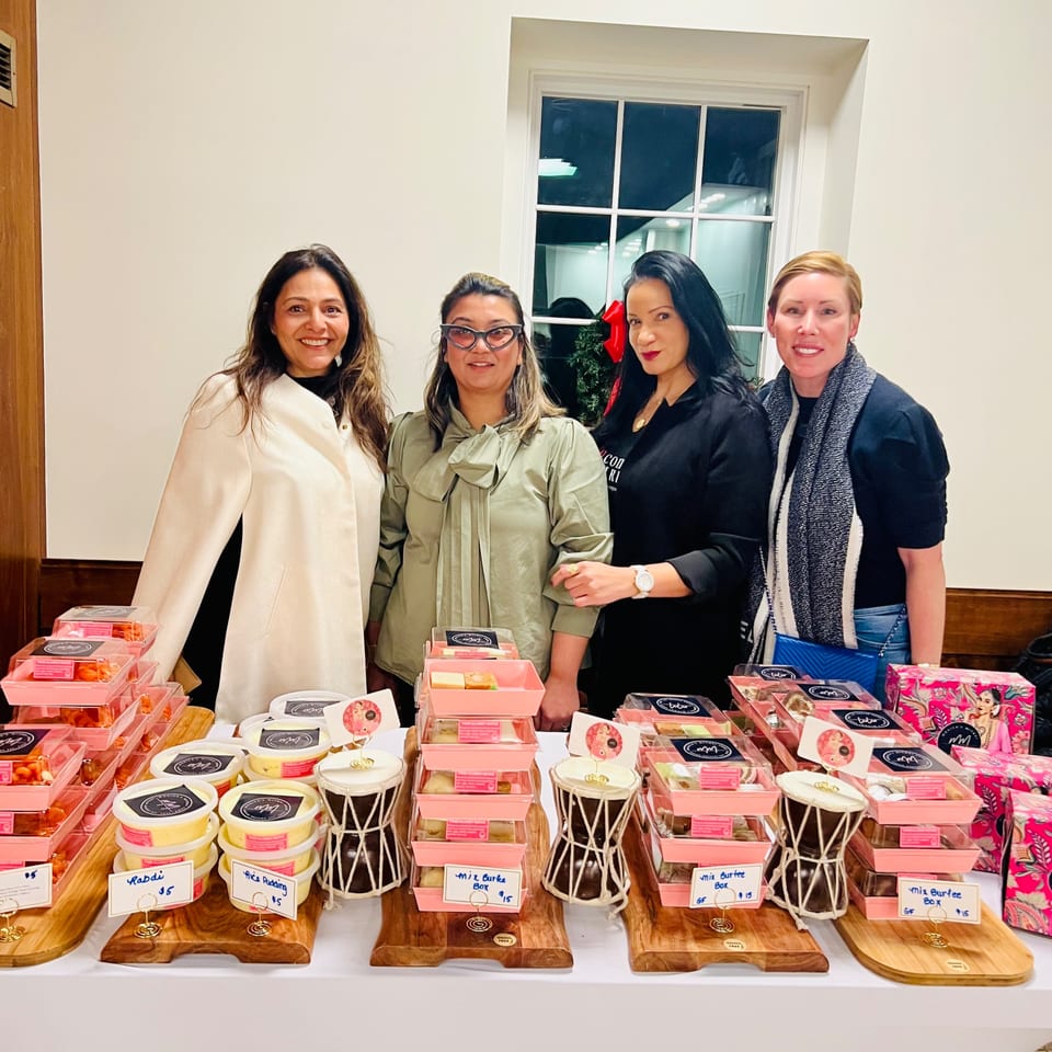 Four women stand behind a table laden with Monica's Mithai Cafe & Bar's Indian sweets and puddings.