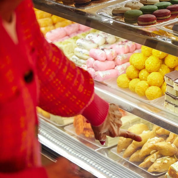 A person in a red sweater points at a display case filled with colorful Indian mithai sweets.