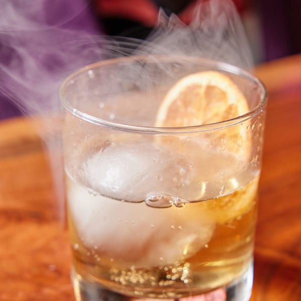 Steaming cocktail with a lemon slice and large ice cubes on a wooden table.