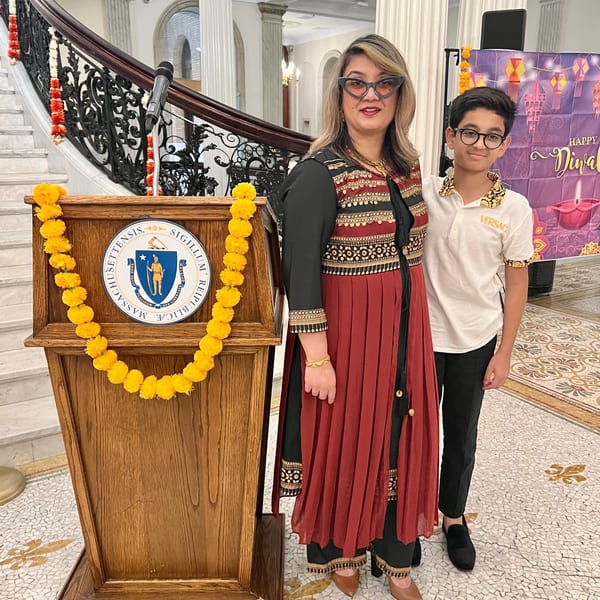 A woman and boy stand by a podium adorned with marigolds, with a Diwali banner in the background.