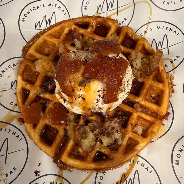 A Monica's Mithai waffle topped with ice cream, syrup, and rose petals on branded paper.