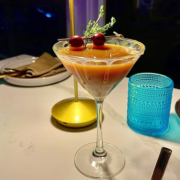 A martini glass filled with a caramel-colored drink, garnished with cranberries and a sprig of thyme.