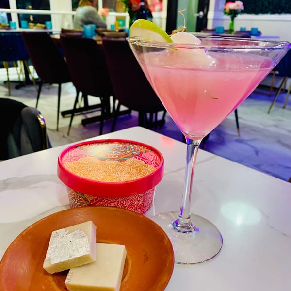 Pink cocktail with lime and lychee, mithai box, and kaju katli on a table at Monica's Mithai Cafe & Bar.