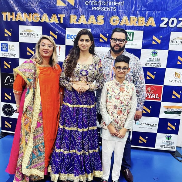 Four people in traditional Indian attire pose in front of a blue and white checkered banner.