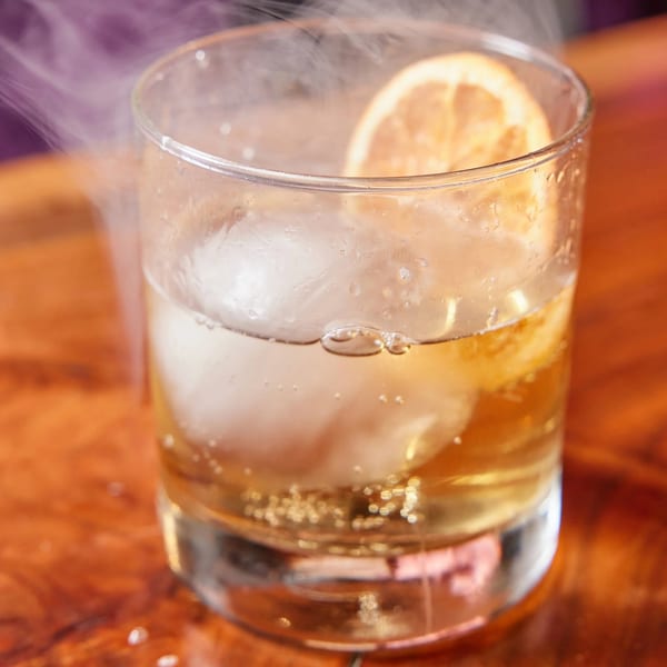 A smoking cocktail with a lemon slice and large ice cube in a glass on a wooden table.