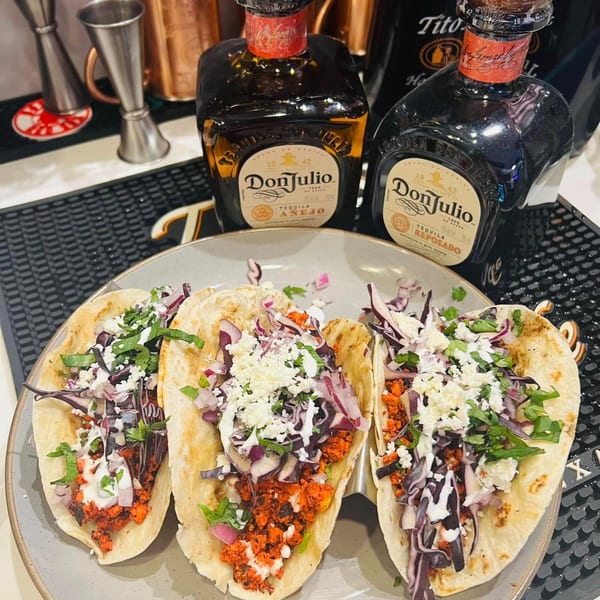Three Indian-style tacos with red filling, purple cabbage, white cheese, and cilantro, with tequila bottles in the background.