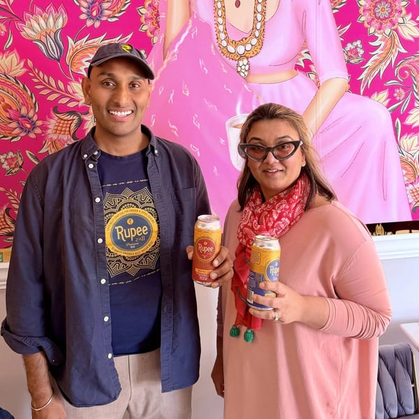Two people smiling and holding Rupee beer cans in front of a vibrant pink floral wall mural.
