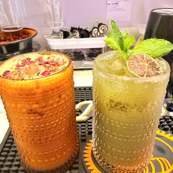 Two Indian cocktails, one orange with rose petals, one green with mint and dried lime, on a bar mat.