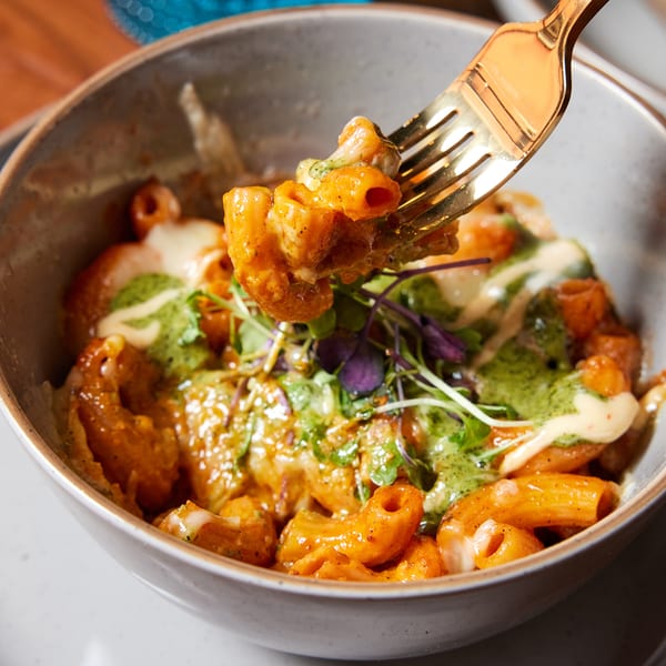 A golden fork lifts a piece of orange pasta from a bowl of Indian-style pasta with green sauce.