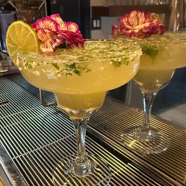 Two margaritas garnished with lime slices and pink flowers sit on a metal bar grate.