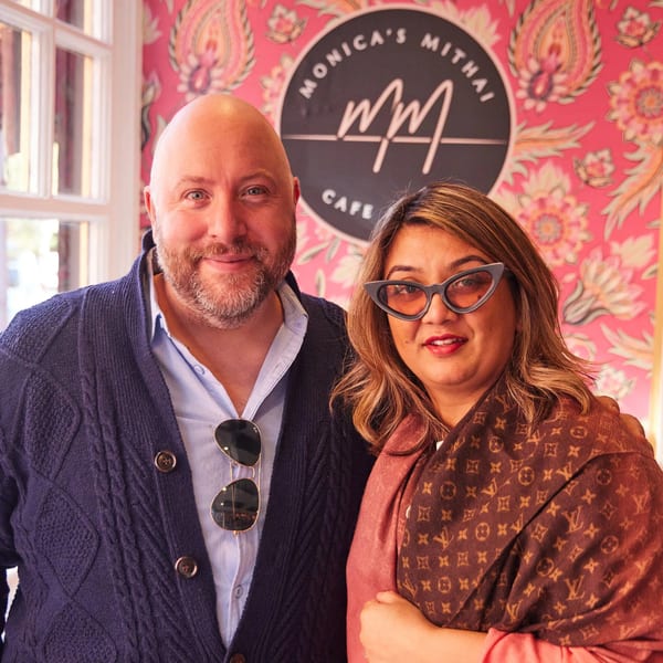 Two people smiling in front of a pink floral wall with the Monica's Mithai Cafe logo.