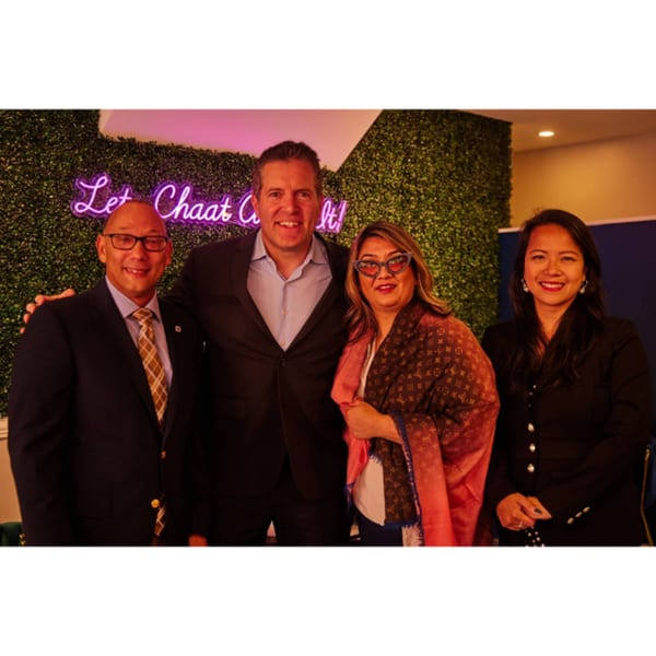 Four people, two men and two women, smiling in front of a green wall with a neon sign.