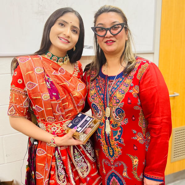 Two women in vibrant Indian attire, one holding a box of Monica's Mithai sweets, smiling at the camera.